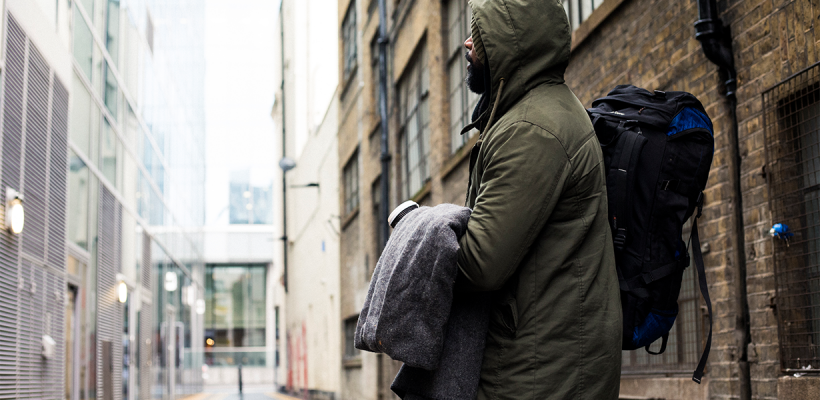 A man in a warm green coat holding a sleeping back, standing in a grey street and looking at the building in front of him.