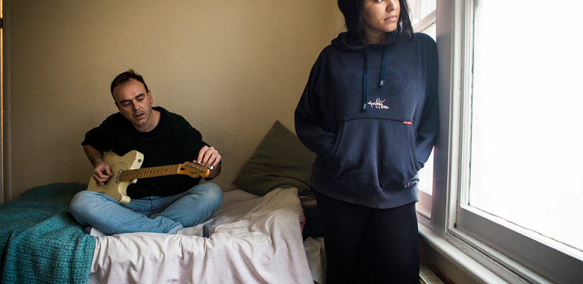 A man with a guitar sitting on a bed. Next to him a woman with a hoodie is looking out of the window.