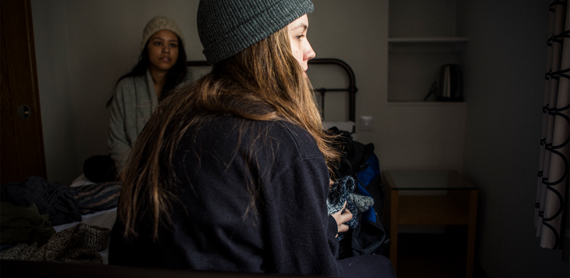 Two girls sitting on a bed in a hostel room, looking out into the distance.