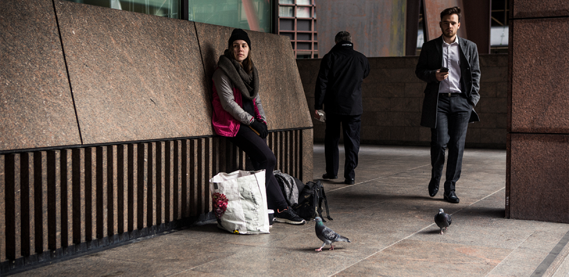 A homeless man leaning behind a wall, while office workers are walking past him.